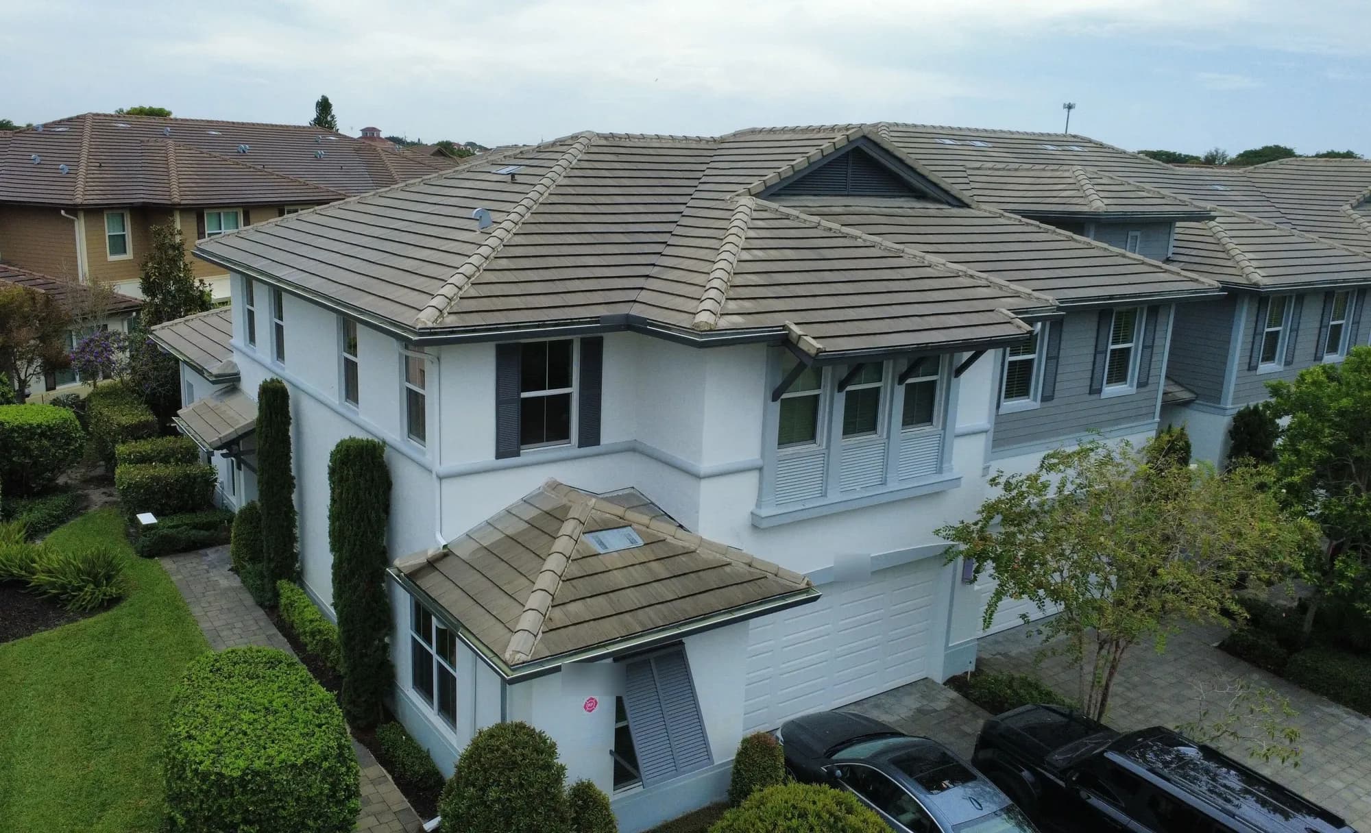 Drone view of a two-story white home with a gray flat-tile hip roof and landscaped yard.