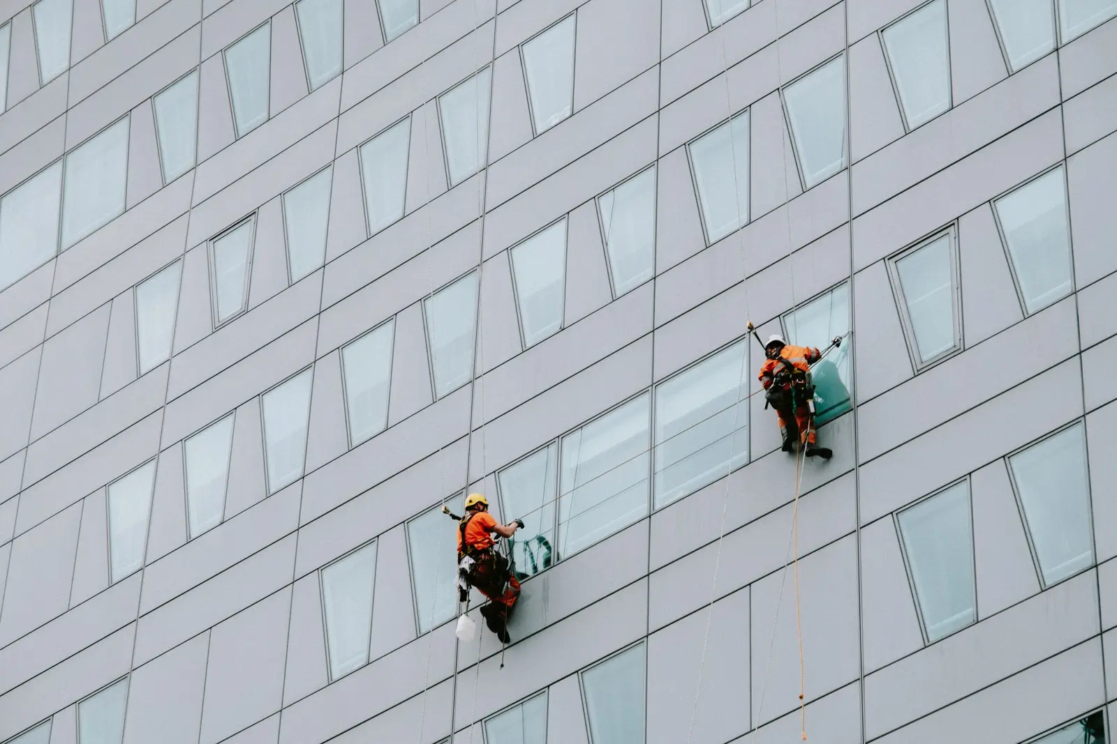 Two rope-access technicians in harnesses and helmets working on a high-rise facade.